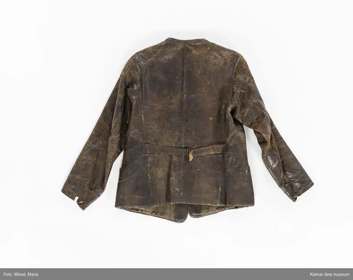 KLM 46056:1. Jacka. Skinnjacka. Arbetsjacka. Använd ytterplagg. Av mörkbrunt skinn. Kort modell. Dubbelknäppning med fem par knappar, alla udda, och knapphål. Utan krage. Dolda fickor fram samt bröstficka på vänster sida. Slejf med spänne i ryggen. Lång ärm som avslutas med kort sprund som knäpps med knapp och knapphål. Grönbrunt foder av kypertvävd bomull/ylle. Fastsydd hank av brun mocka i fodret i nacken.