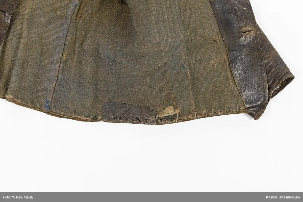 KLM 46056:1. Jacka. Skinnjacka. Arbetsjacka. Använd ytterplagg. Av mörkbrunt skinn. Kort modell. Dubbelknäppning med fem par knappar, alla udda, och knapphål. Utan krage. Dolda fickor fram samt bröstficka på vänster sida. Slejf med spänne i ryggen. Lång ärm som avslutas med kort sprund som knäpps med knapp och knapphål. Grönbrunt foder av kypertvävd bomull/ylle. Fastsydd hank av brun mocka i fodret i nacken.