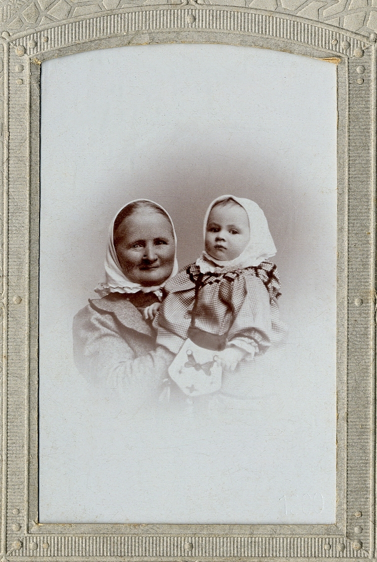 En medelålders kvinna i sjalett, som håller upp en liten flicka i rutig kolt och sjalett. I nedre högra hörnet 
syns inpräglat årtal: "1899". 
Bröstbild, en face. Ateljéfoto. 

Fotografens svärmor och dotter.