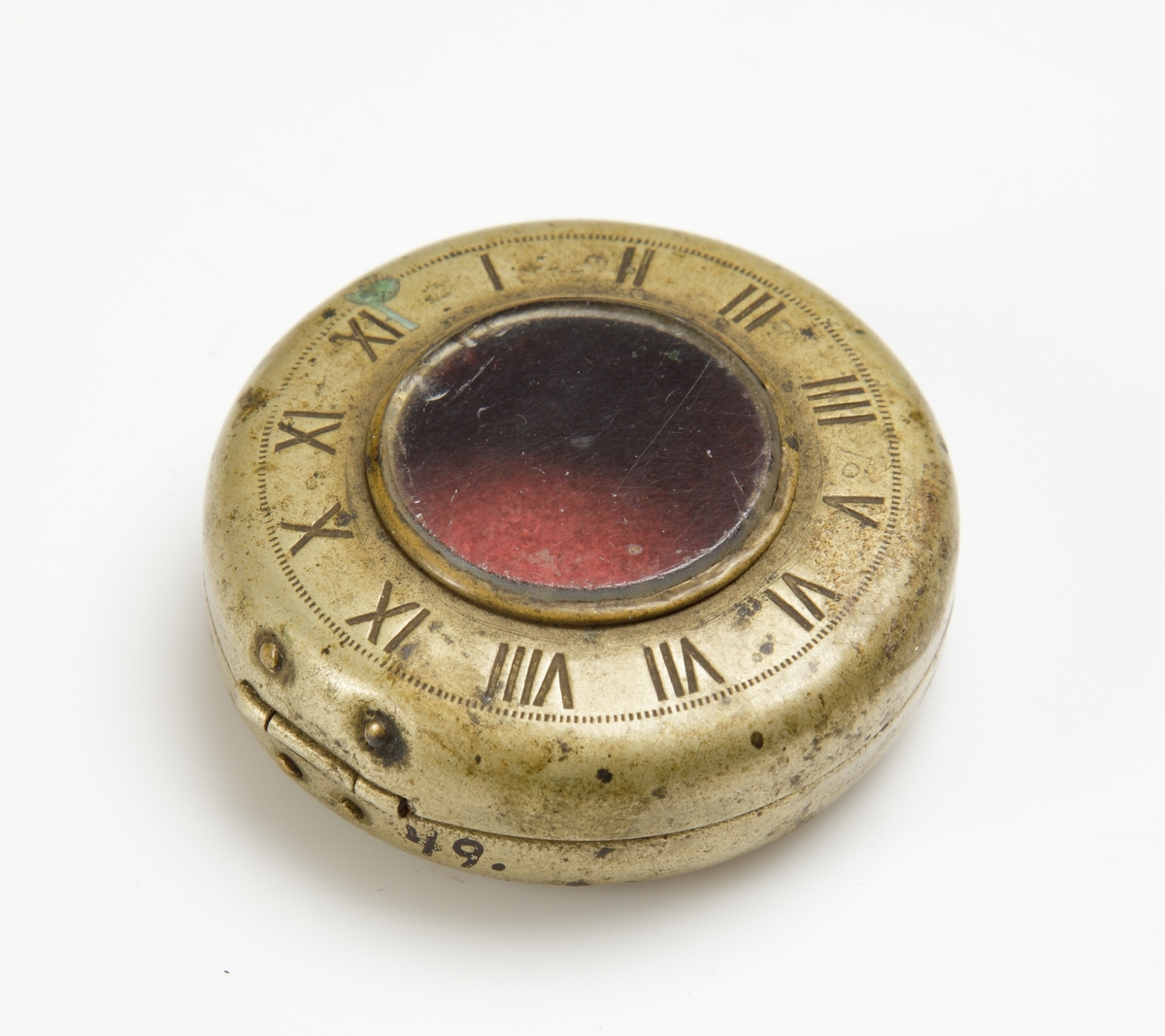 Rund mässingsboett. På locket urtavla med glas i mitten, omkring detta romerska siffror. Stämplat 20½. 
Röd tygbit inuti.