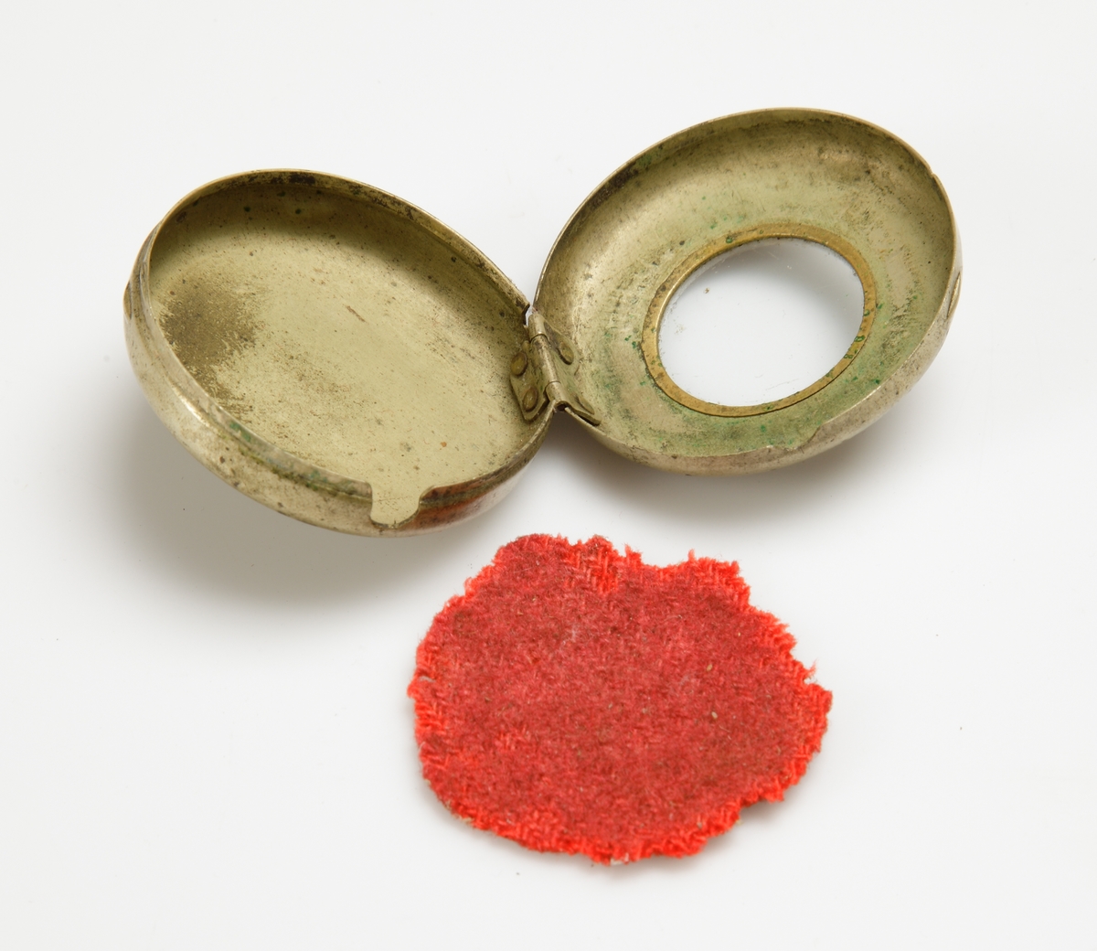 Rund mässingsboett. På locket urtavla med glas i mitten, omkring detta romerska siffror. Stämplat 20½. 
Röd tygbit inuti.