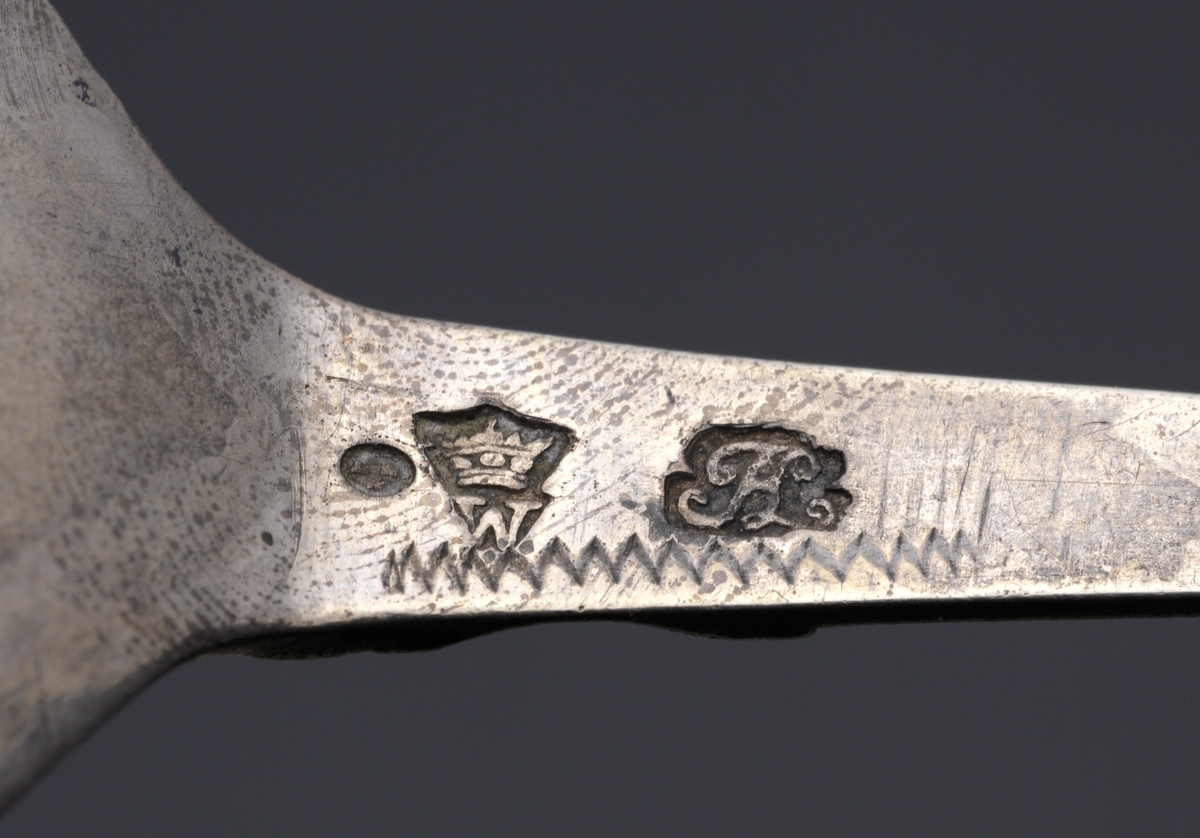 Brännvinssked av silver. 
S.k. kronsked med nästan runt skedblad, kort platt skaft, dekorerat med tvärräfflor längst ner. Skaftknopp i form av krona, 
sammansatt av fyra gotiska korsblommor. I kronans hörn hänger spiralvridna ringar (två saknas). 
Bladet har dekor i form av ett förgyllt kors med en graverad blomma i mitten. Även inre kanten av bladet är förgyllt. 
Stämplar och s.k. åldermansranka på skaftets baksida.

Inskrivet i huvudbok 1867.
