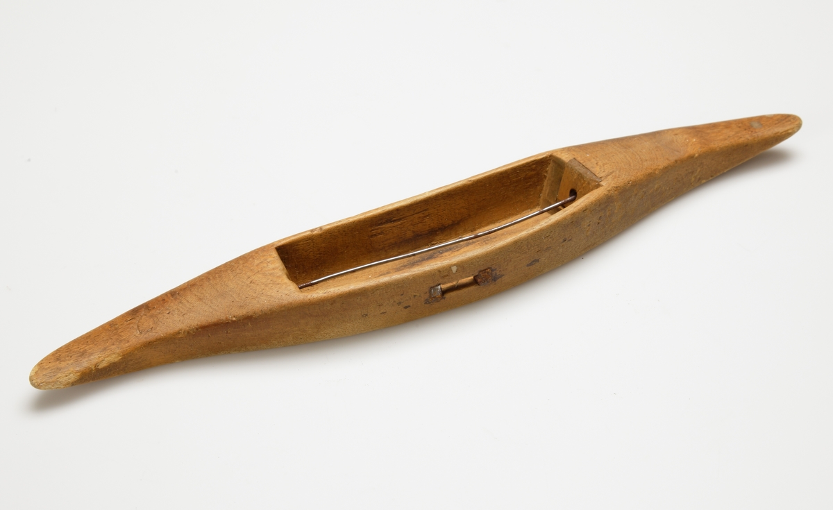 Vävskyttel av trä (8 st).
Vågformad kontur. Avlångt hål med små benplattor omkring hålen för stången. 
a) med svart tråd
f) med tom tråd "tuta". 1894 skrivet med bläck.
Resterande skyttlar är tomma.

Mått:
Längd: 280 mm
Längd hål: 90-105 mm
Bredd: 35-45 mm