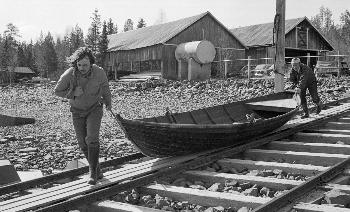 Tømmerfløterne Håvard Fjeld og Sverre Nyhus drar ut et av robåtene som skulle brukes under fløtinga på Osensjøen i grensetraktene mellom Trysil og Åmot kommuner i Hedmark i 1984. Fotografiet er fra Glomma fellesfløtingsforenings anlegg ved Sørlistøa på vestsida av innsjøen, der materiellet som ble brukt i denne virksomheten ble vinterlagret. I bakhgrunnen på dette fotografiet ser vi to parallelle, lange bindingsverkshus. Det som lå nærmest innsjøen var ved- og lensehuset, som har ei grunnflate på 226 kvadratmeter. Bakenfor lå båthuset, med 198 kvadratmeters grunnflate, hvor alle de mindre farkostene som ble brukt under fløtinga ble lagret mellom driftssesongene. Fra den nordvestre gavlen på båthuset gikk det en skinnegang som svingte mot slippen for slepebåten Trysilknut og ned mot sjøen. Da denne robåten ble sjøsatt brukte ikke karene den skinnegående vogna, men de dro og skjøv båten nedover mot vannet med kjølen på bord de hadde lagt på svillene mellom skinnene. Osensjø-fløterne disponerte 5-6 cirka 5 meter lange robåter, tre varpebåter av stål og en stor slepebåt (Trysilknut), som også hadde stålskrog. I tillegg fantes det en inspeksjonsbåt av stål, «Ener», som fløtersjefen disponerte. Alle stålbåtene hadde dieselmotorer. Diesellageret var den digre, sylindriske ståltanken vi ser framfor gavlveggen på ved- og lensehuset i bakgrunnen.