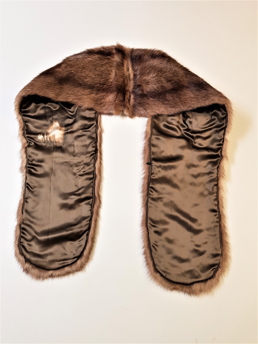 Midjelang stola, som kan lukkes med én kraftig hekte midt foran. Ved halsåpningen er skinnet splittet, slik at det fremkommer en effekt av et legg rundt nakken, med en ytre del som rager litt høyere enn det smalere stykket innerst mot halsen. Øvre del har søm midt bak, og skinnet er brettet inn mot nakken. På grunn av splitten som er skåret på hver side av nakken, dannes det et skrått skulderparti. Nedenfor skuldrene er det skjøtt på lange, jevnt brede og avrundede forstykker.  Plagget er fôret, og har en stor fuktskjold på den ene siden. Fôret er av blank, myk, silkeaktig sateng, men kan kanskje være sekundært og av syntetfiber. Ingen spor etter etikett. Pelsverket er fra dyrets ryggstykke, og er anvendt slik at den mørke stripen fra ryggstykket danner et dekorativt element.