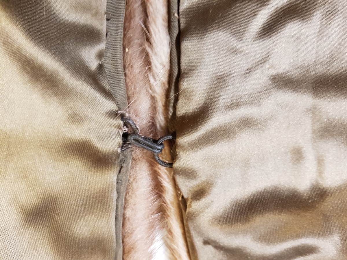 Midjelang stola, som kan lukkes med én kraftig hekte midt foran. Ved halsåpningen er skinnet splittet, slik at det fremkommer en effekt av et legg rundt nakken, med en ytre del som rager litt høyere enn det smalere stykket innerst mot halsen. Øvre del har søm midt bak, og skinnet er brettet inn mot nakken. På grunn av splitten som er skåret på hver side av nakken, dannes det et skrått skulderparti. Nedenfor skuldrene er det skjøtt på lange, jevnt brede og avrundede forstykker.  Plagget er fôret, og har en stor fuktskjold på den ene siden. Fôret er av blank, myk, silkeaktig sateng, men kan kanskje være sekundært og av syntetfiber. Ingen spor etter etikett. Pelsverket er fra dyrets ryggstykke, og er anvendt slik at den mørke stripen fra ryggstykket danner et dekorativt element.