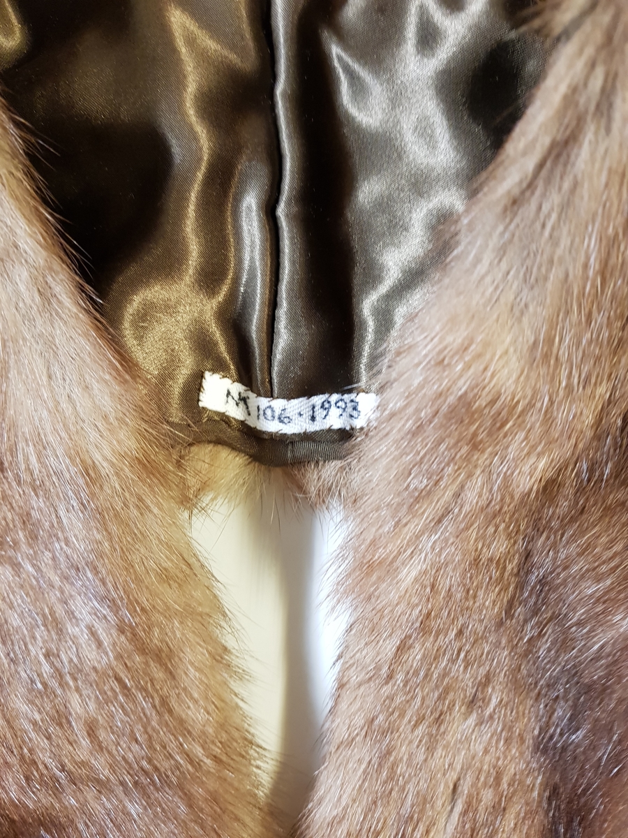 Midjelang stola, som kan lukkes med én kraftig hekte midt foran. Ved halsåpningen er skinnet splittet, slik at det fremkommer en effekt av et legg rundt nakken, med en ytre del som rager litt høyere enn det smalere stykket innerst mot halsen. Øvre del har søm midt bak, og skinnet er brettet inn mot nakken. På grunn av splitten som er skåret på hver side av nakken, dannes det et skrått skulderparti. Nedenfor skuldrene er det skjøtt på lange, jevnt brede og avrundede forstykker.  Plagget er fôret, og har en stor fuktskjold på den ene siden. Fôret er av blank, myk, silkeaktig sateng, men kan kanskje være sekundært og av syntetfiber. Ingen spor etter etikett. Pelsverket er fra dyrets ryggstykke, og er anvendt slik at den mørke stripen fra ryggstykket danner et dekorativt element.