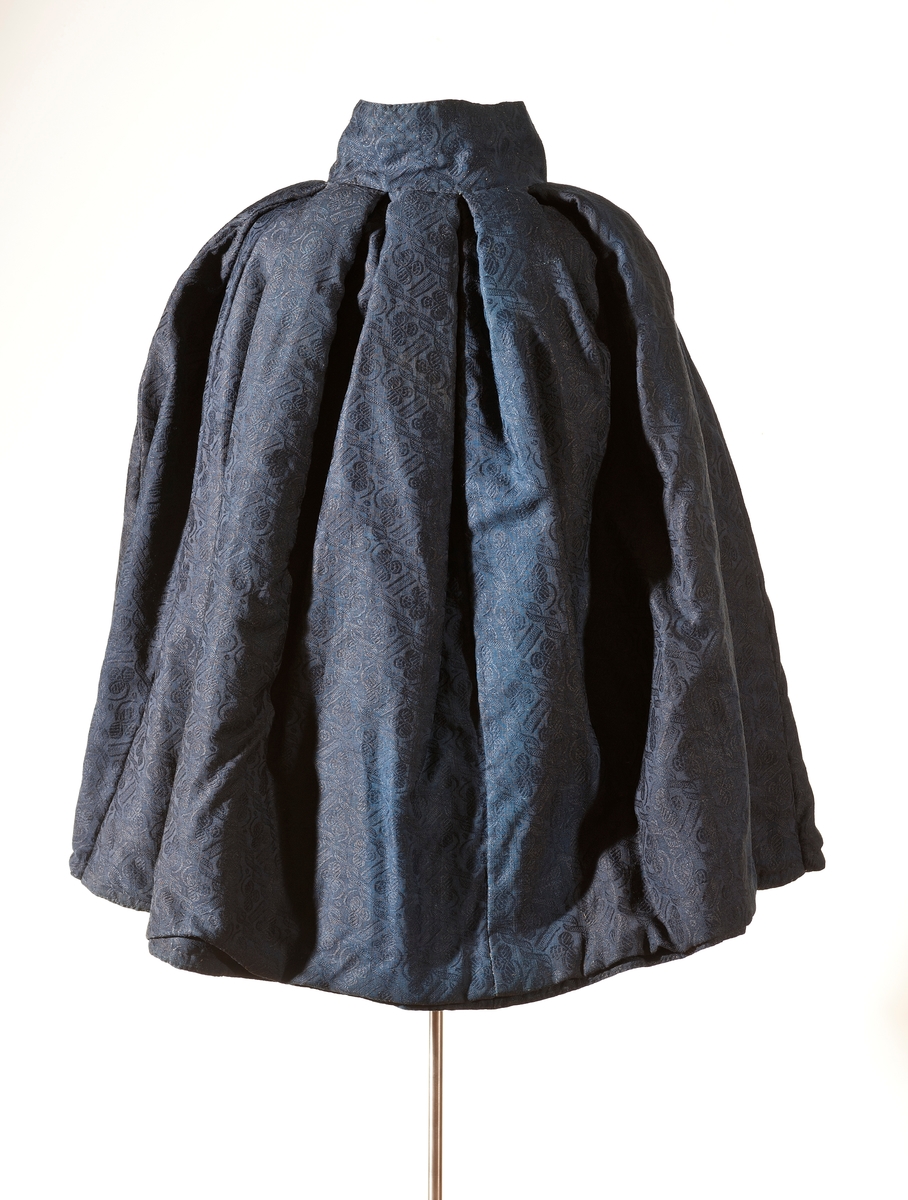 Mørk blå kep. Ytterstoff i everlasting. Fôr i hjemmevevet ullstoff i kypert. Halslinning er fôret med kattun (mørk brunt med blomster i rosa/blått). Kepen lukkes med ripsvevd knytebånd i halsen. 