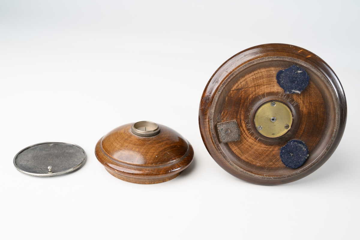Avmagnetiseringsapparat för borttagande av magnetism i klocka. Apparaten består av en rund, svarvad behållare med lock. Under locket finns en bricka av metallnät, där objekt som ska avmagnetiseras placeras. Avmagnetiseringsmekaniken under bricka är av mässing. På sidan av träbehållaren sitter en mässingsskruv som har kontakt med insidans utrustning. Lodrät spricka i träbehållaren.