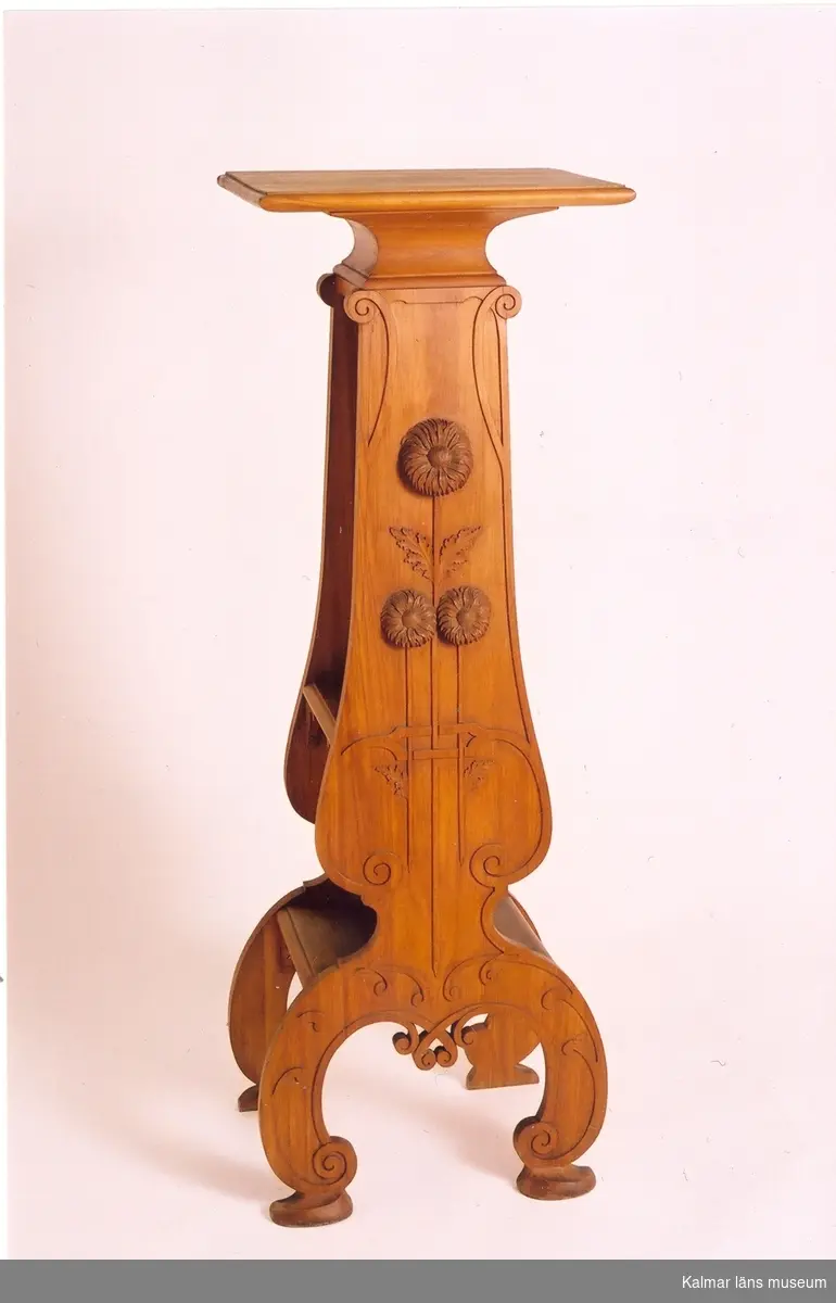 KLM 25247. Piedestal. Av plommonträ. Sidostöden med skuren ornamentik i form av naturalistiska blommor, krysantemum i hög relief. Mellan stöden två hyllplan och överst en platta. Signerad: Anton Wahledow 1899 Kalmar.