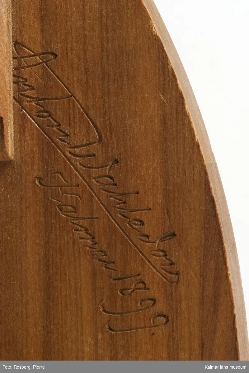 KLM 25247. Piedestal. Av plommonträ. Sidostöden med skuren ornamentik i form av naturalistiska blommor, krysantemum i hög relief. Mellan stöden två hyllplan och överst en platta. Signerad: Anton Wahledow 1899 Kalmar.