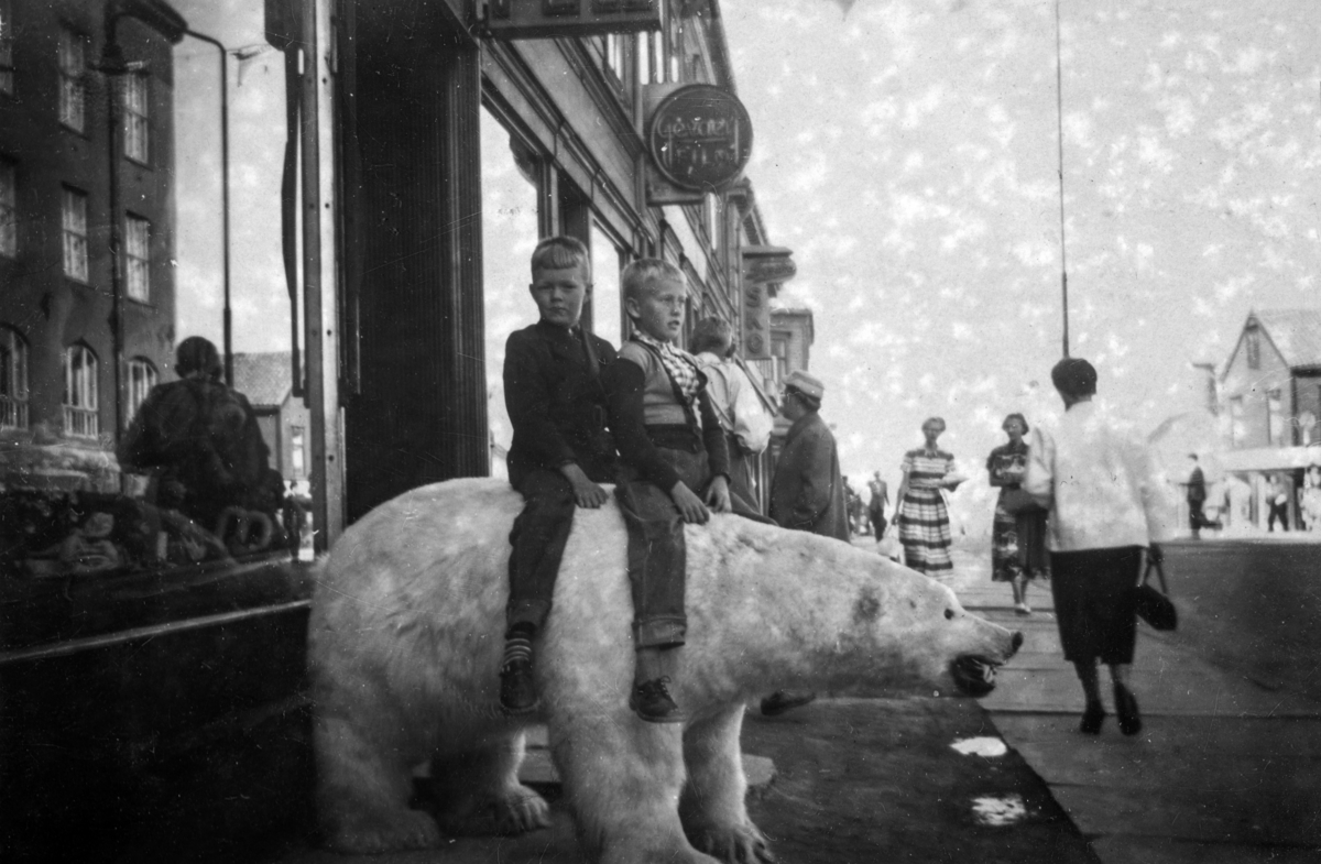 To gutter sittende på en utstoppet isbjørn i Tromsø. Guttene på isbjørnen er fra venstre Trond Arve Nilsen og Åge Thomassen, begge født i 1947, og er 8 år når bilde er tatt. Guttenes fedre er begge i fra Alta, men ble evakuert sør-over under krigen. Og ble etter hvert boende i Ottestad sammen med sine familier. Bildet er tatt under en ferie tur i 1956. De reiste med båt i fra Mosjøen til Alta, "Mosjøruta". Reisen fra Ottestad til Alta tok 3 dager. Guttenes fedre er Reidar Nilsen (1919-1990 og Rolf Thomassen (1914-1989) begge født og oppvokst i Alta.