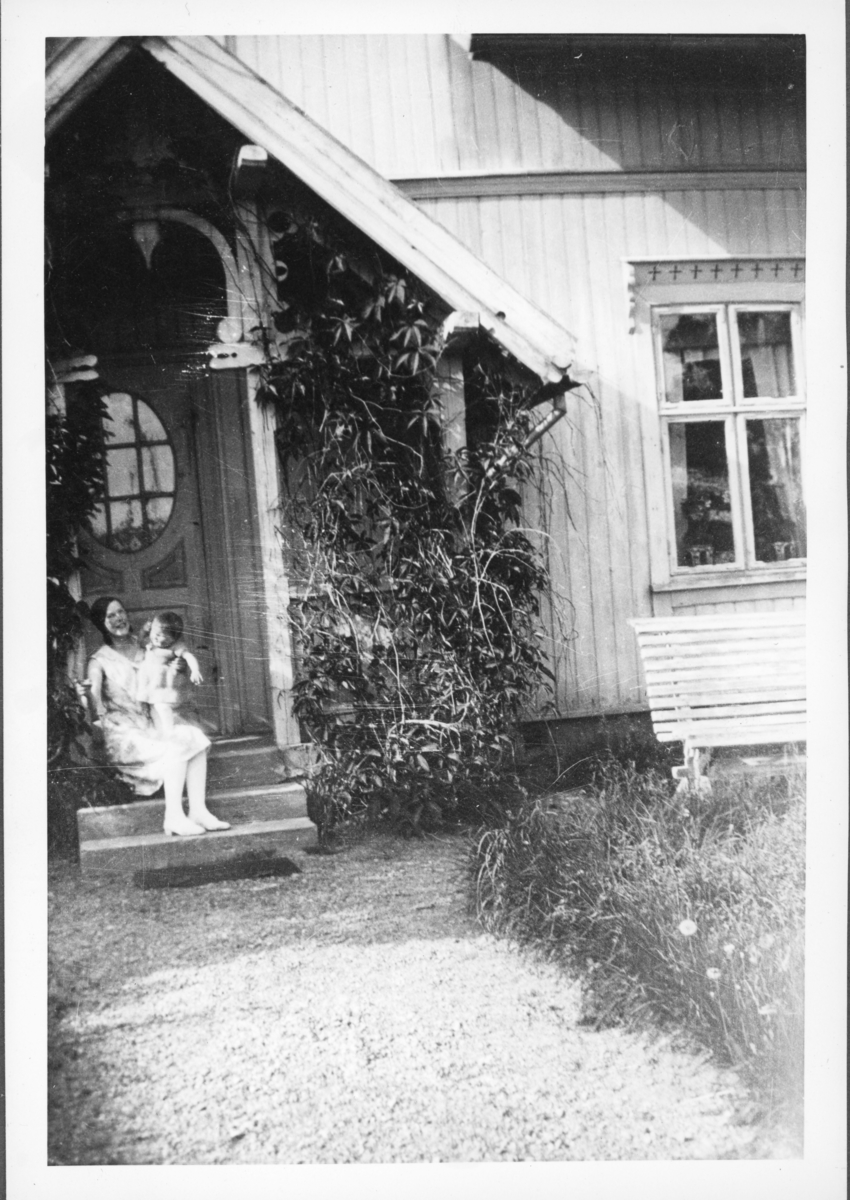Kvinne med barn på fanget foran inngangsdør. Trolig 1930-tall.