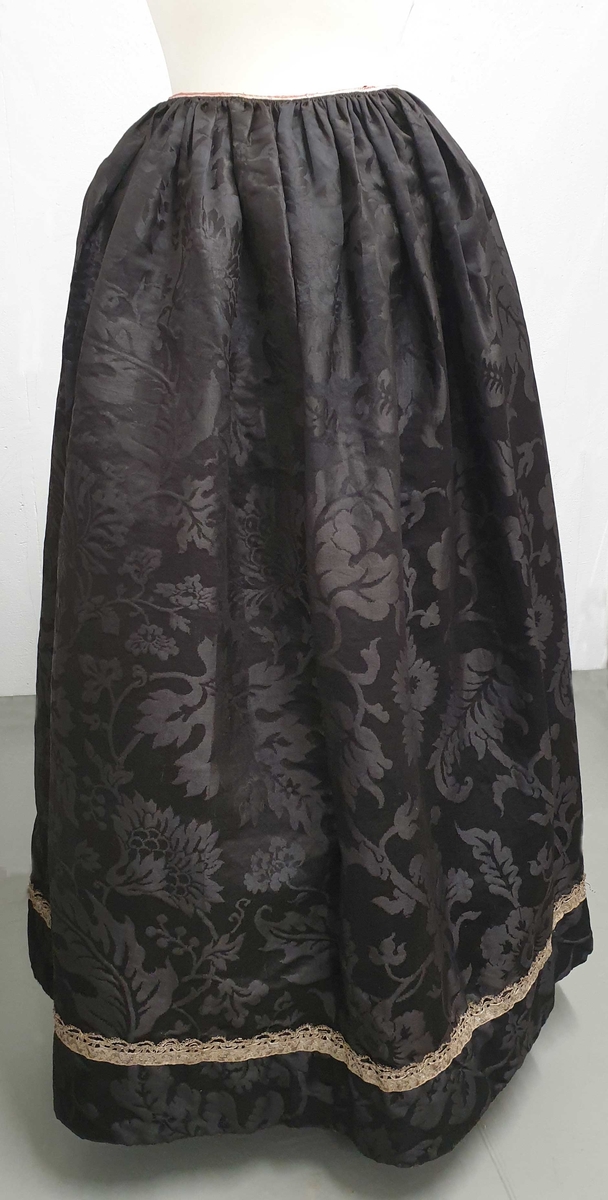 Forkle til brudedrakta. Av svart silkedamask med bord i sølvknipling, og med knytting i livet.