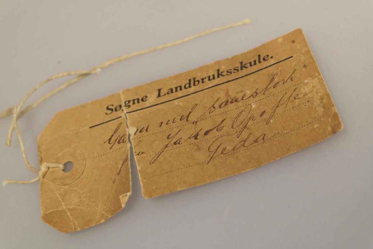 Sauestokk i tre. Stokken er buet og har en rund utsparing på midten samt to små hull på hver side av utsparingen. På den ene siden er det inngravert ett kryss samt et håndskrevet personnavn. En merkelapp med håndskrevet tekst har vært festet til stokken med hyssing. Stokken ble brukt for å hindre sau å hoppe over gjerde o.l. ved å feste den til sauens ben.