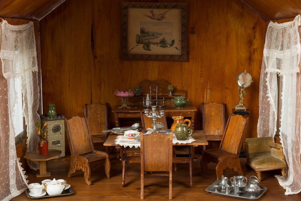 Dukkehus i tre. Huset har 3 etasjer og et spisstak. Hver etasje har 2 vinduer med gardiner. Overflaten er beiset og lakkert.