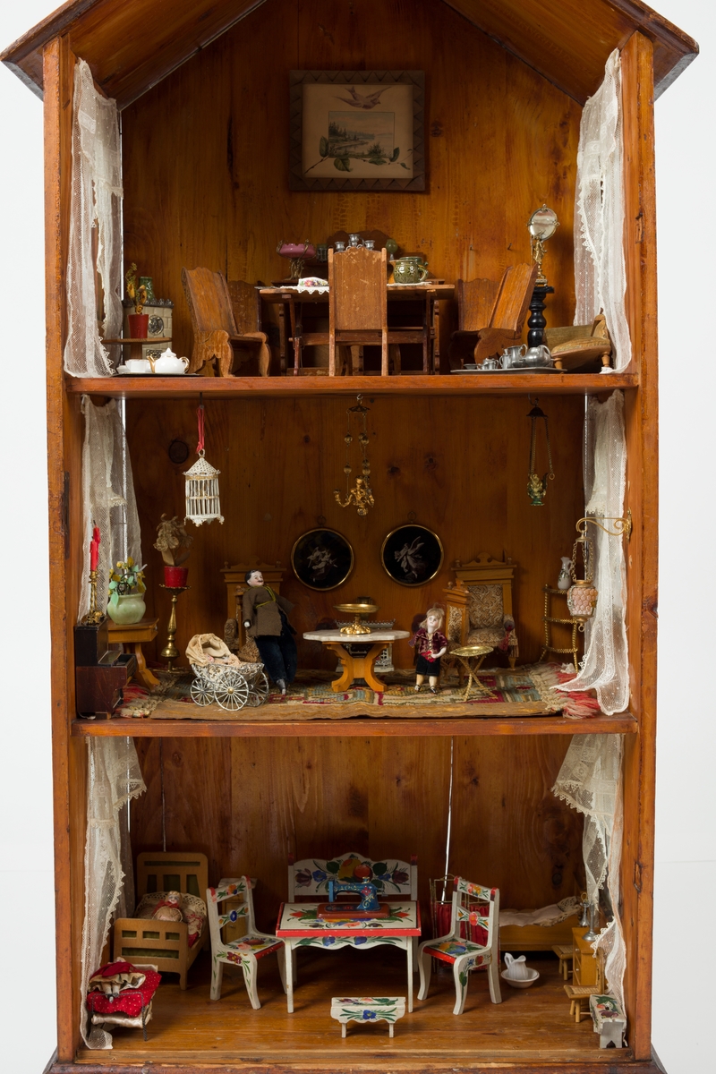 Dukkehus i tre. Huset har 3 etasjer og et spisstak. Hver etasje har 2 vinduer med gardiner. Overflaten er beiset og lakkert.