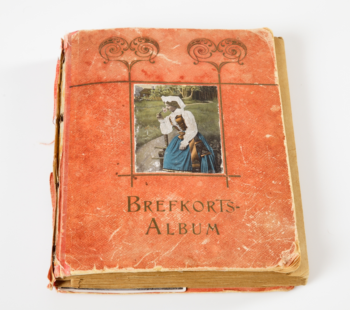 Fotoalbum med röda pärmar. Framsidan med en kolorerad bild av en kvinna som luktar på en blomma. Nedanför bilden är "Brefkortsalbum" skrivet. Runt bilden en ram med två snirkliga hjärtan ovanför.