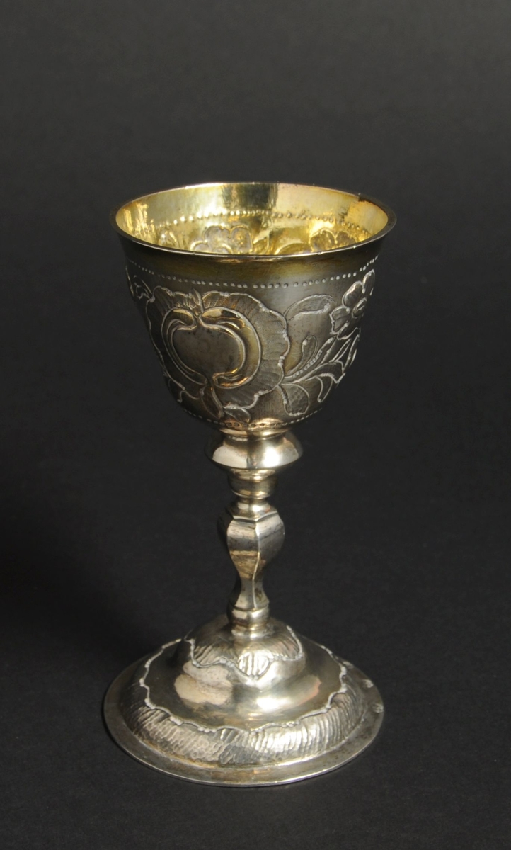 Beger av sølv. Begeret har stett og rund sokkel. På klokken er det to border med dekorative punslinger, mellom bordene er det drevet dekor av blomster. Siselert bølgedeker på sokkel.