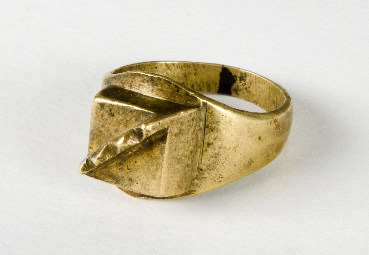 Fingerring av metall, sannolikt mässing. Lödning i "bakkant". Dekoren är en kvadrat med en upphöjd "kam" med fyra taggar. En trolig datering är historisk tid.
