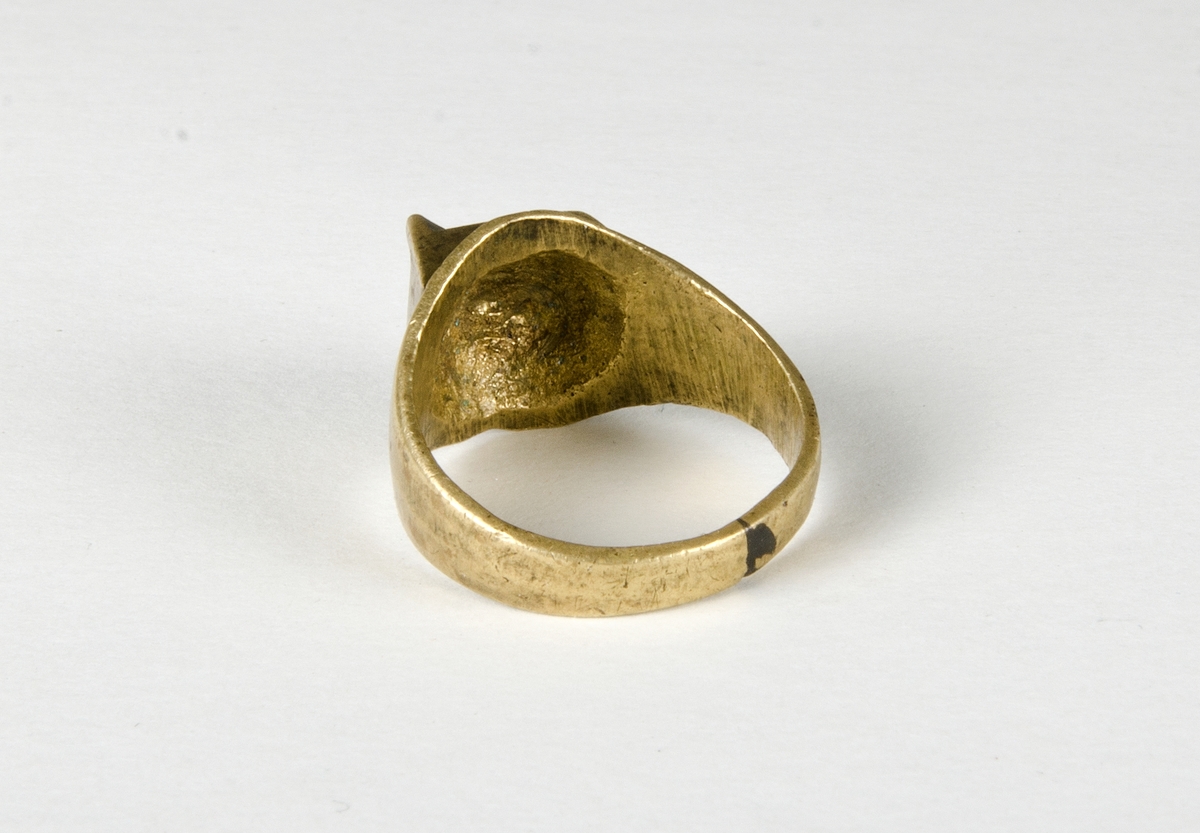 Fingerring av metall, sannolikt mässing. Lödning i "bakkant". Dekoren är en kvadrat med en upphöjd "kam" med fyra taggar. En trolig datering är historisk tid.