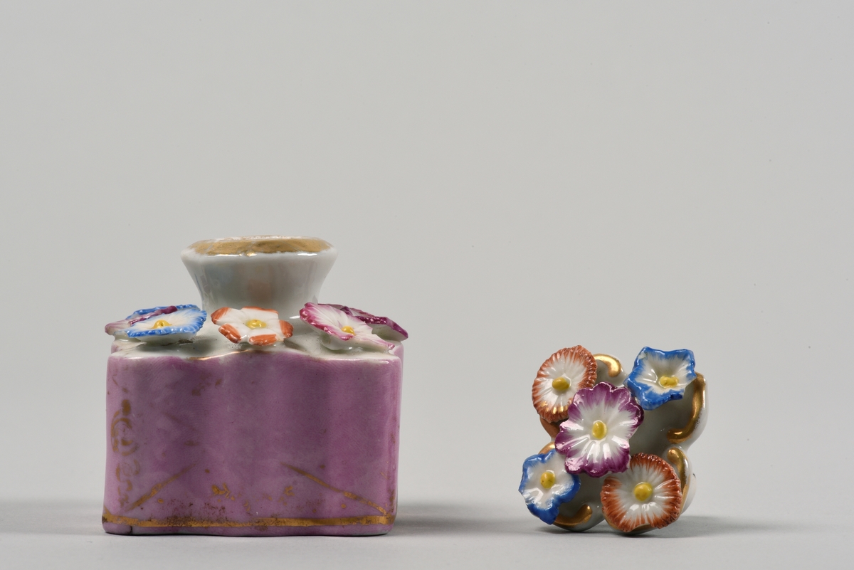 En av två parfymplaskor av porslin. Vågigt utformad fyrsidig flaska med kort hals. På ovansidan dekorerad med plastiskt utformade blommor, åtta stycken i olika färger. Sidorna är rosa med en guldrand nära botten. Proppen är i form av ett fyrpassat löv med fem plastiskt formade blommor, i olika färger.