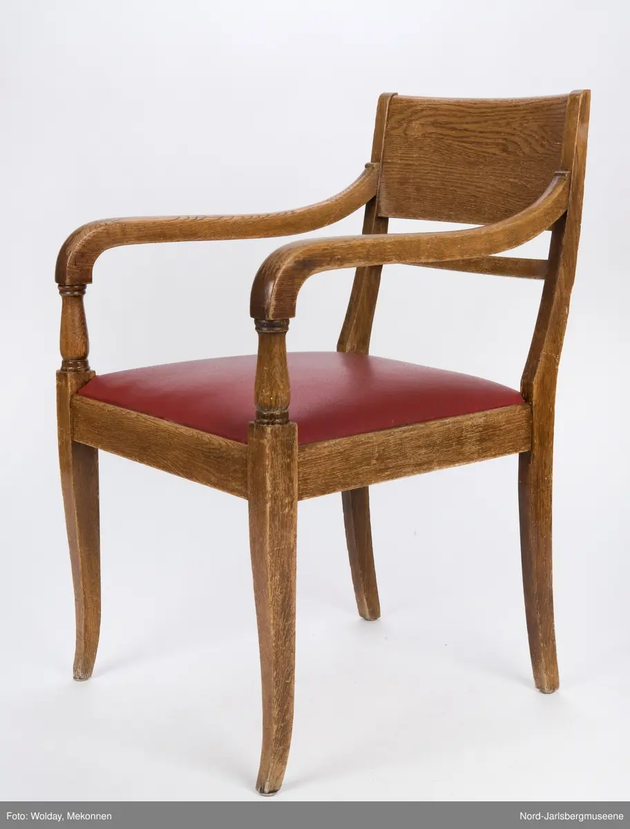 Stol i heltre med stoppet sete med rødt trekk. Stolen har forseggjorte armlener som avsluttes i en myk bue, med dreide detaljer. Ryggstø har en bred plate øverst og en tynnere list under den.