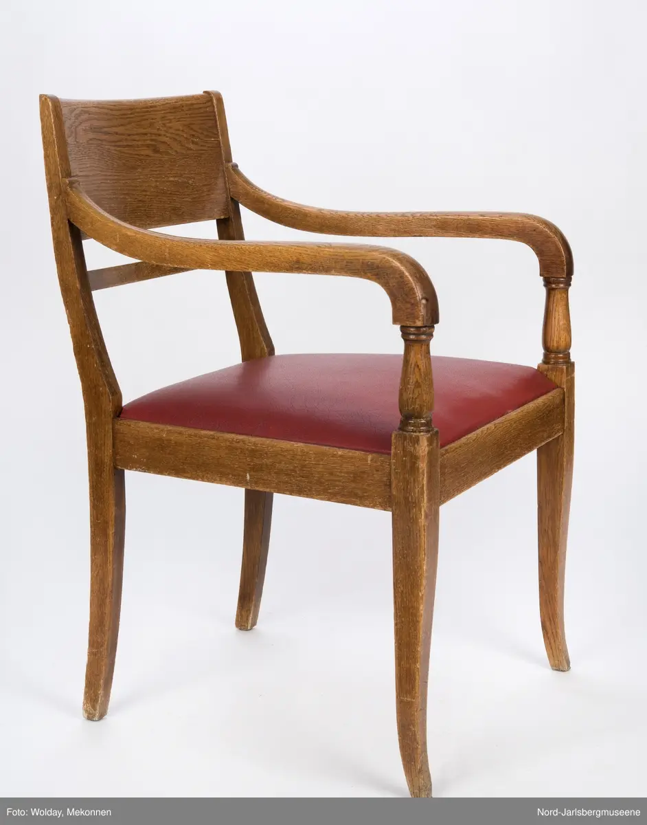 Stol i heltre med stoppet sete med rødt trekk. Stolen har forseggjorte armlener som avsluttes i en myk bue, med dreide detaljer. Ryggstø har en bred plate øverst og en tynnere list under den.