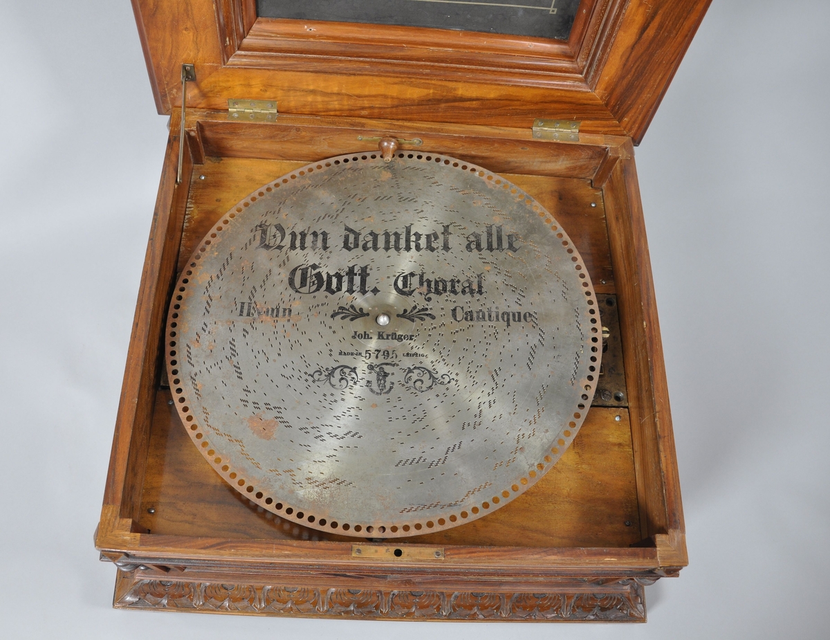 Spilledåse av tre. Spilledåsen har rektangulær form. Står på sokkel med fire bein. Øverste del av sokkel har en borde med palmetter, denne delen har en profilert svai. Selve kassen har dreide pillarer i hjørnene med dekorative kanneleringer. På sidene av kassen er det plater med valbjørk. På framsiden av kassen er det en dekorativ, konsentrisk rombe. Lokket er profilert, med oppstånde midtdel. Midtdelen har svungne sider, med flatskurd av tynne ranker. På toppen av midtdelen, er det intarsia med motiv av lyse blomster på mørk bakgrunn. Treverket er lakkert. 
Lokket er hengslet, det er et litografi/trykk på innside av lokk med motiv av barn som spiller instrumenter.