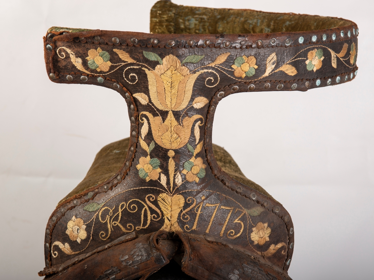 Brodert lær. Grønn mønstret  fløyel i setet og på armlene. Messinghode på fremre del av rygglenet. Initialene KDS og årstallet1773 brodert på. Løs narv på kappene, redrot.