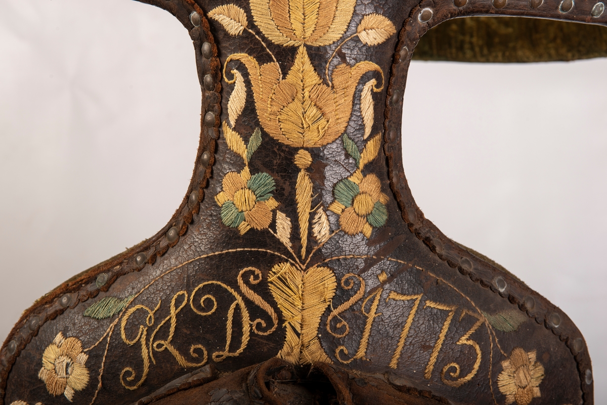 Brodert lær. Grønn mønstret  fløyel i setet og på armlene. Messinghode på fremre del av rygglenet. Initialene KDS og årstallet1773 brodert på. Løs narv på kappene, redrot.