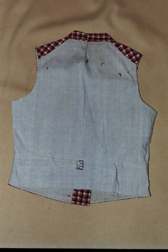 Vest i rutet bomullslerret i fargene rødt, olivengrønt/grønnnbrunt og ubleiket hvitt. Fôr og rygg i linlerret. Vesten er dobbeltspent med svarte, stofftrukne knapper. Den har sjalskrage og spensel i ryggen. Plagget er helt håndsydd. På forstykkene er det to lett skråstilte lommer og vesten buer ned midt foran.