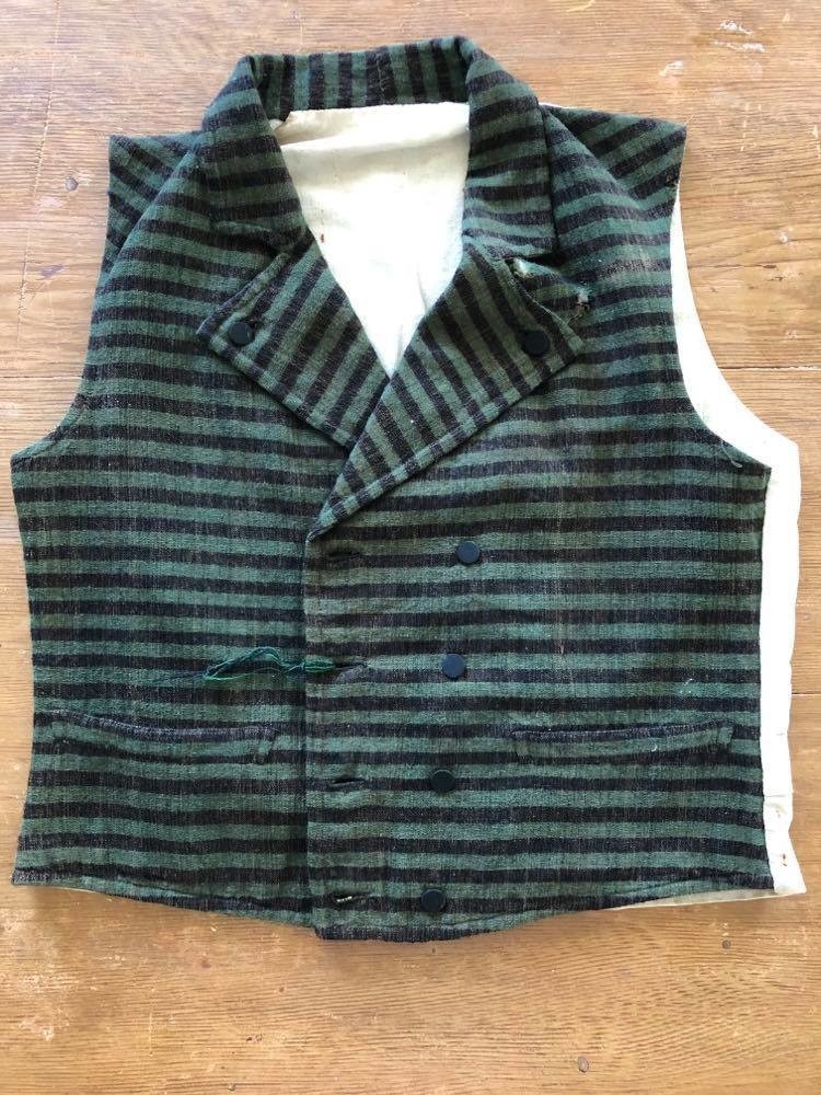 Dobbeltspent vest i ullverken, stripet i grønt og svart (sauesvart) med brun renning. Dobbel rygg i bomullslerret. Halvlinslerret (bomull/stry) i fôret i framstykkene. Vesten har fire knapphull sydd med kamgarnsullgarn og svarte knapper trukket med svart bomullssateng med brun renning. Plagget har nedbrettet krage og slag/reverser som kan kneppes mot bolen. Knapp og knapphull. Vesten buer ned midt framme og har svakt skråstilte lommer. Vattering av stry i framstykkene. I ryggen buer vesten opp midt bak. Den er vrangsydd langs ytterkantene og stukket ned med maskin, 0,8 cm fra kanten. (Registrering ved Bunad- og folkedraktrådet/Norsk institutt for bunad og folkedrakt v/Jon Fredrik Skauge, 2005, revidert v/Bjørn Sverre Hol Haugen, 2020.)