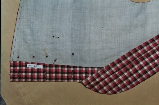 Vest i rutet bomullslerret i fargene rødt, olivengrønt/grønnnbrunt og ubleiket hvitt. Fôr og rygg i linlerret. Vesten er dobbeltspent med svarte, stofftrukne knapper. Den har sjalskrage og spensel i ryggen. Plagget er helt håndsydd. På forstykkene er det to lett skråstilte lommer og vesten buer ned midt foran.