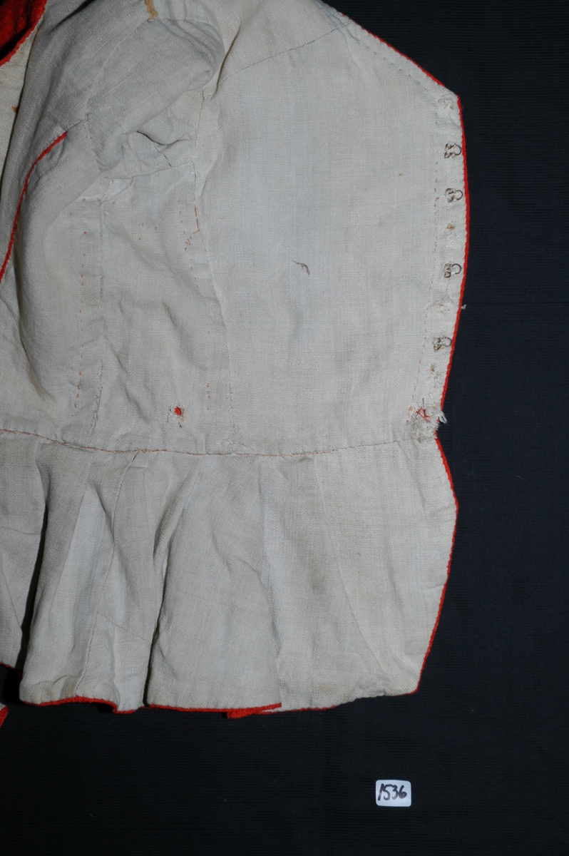 Trøye av rødt ullstoff med linfôr. Stoffet er av kamgarn og ble antageligvis solgt under betegnelsen everlasting på 1700-tallet. Produksjonssted er antageligvis England. Stoffet er i toskaft binding med flotteringer over tre tråder som gir mønstereffekt. Trøya har vært lukket med ni hekter og maljer midt framme. Snitt: Framstykkene er avdelt i midjen med kappe. Ryggstykkene går helt ned i skjøten. Kappa er foldelagt med sju folder (fra 10 cm midt framme). Foldene er 3-3,5 cm brede og 1,5 cm dype. Bua ryggsømmer sydd med spilesøm. Ryggsømmen midt bak er sydd med attersting hver for seg, men festet sammen i fôr og hovedstoff på noen steder. Det er to innsnitt under hvert herme. Ytterkantene er faldet med faldeprikkesting. Det er ekstra prikkestingsrad ved halsringing (1 cm fra ytterkant) og ved hektestillinga omlag 2 cm fra ytterkanten framme. Kappa er sydd til bolen med attersting (tre lag stoff). Fôret i bolen er bretta og lagt til og sydd ned i fôret til kappa. Kappa skrår bakover midt framme. Trange ermer sydd sammen med attersting. Sømmonnet er lagt mot bolen og fôret i bolen er lagt mot ermet og sydd til med faldesting. Skinnhemper til opphenging. Ensømserme med albuestnitt. På unerermene er det et innsnitt. Underermssømmer er sydd med attersting, stoff og fôr hver for seg (festet sammen på enkelte steder). Innsnittene er sydd med stoff og fôr sammen og lagt til en side og stukket ned, faldet til side. (Fra regsitrering ved Bunad- og folkedraktrådet/Norsk institutt for bunad og folkedrakt v/Jon Fredrik Skauge i 2005, revidert v/Bjørn Sverre Hol Haugen 2020). 