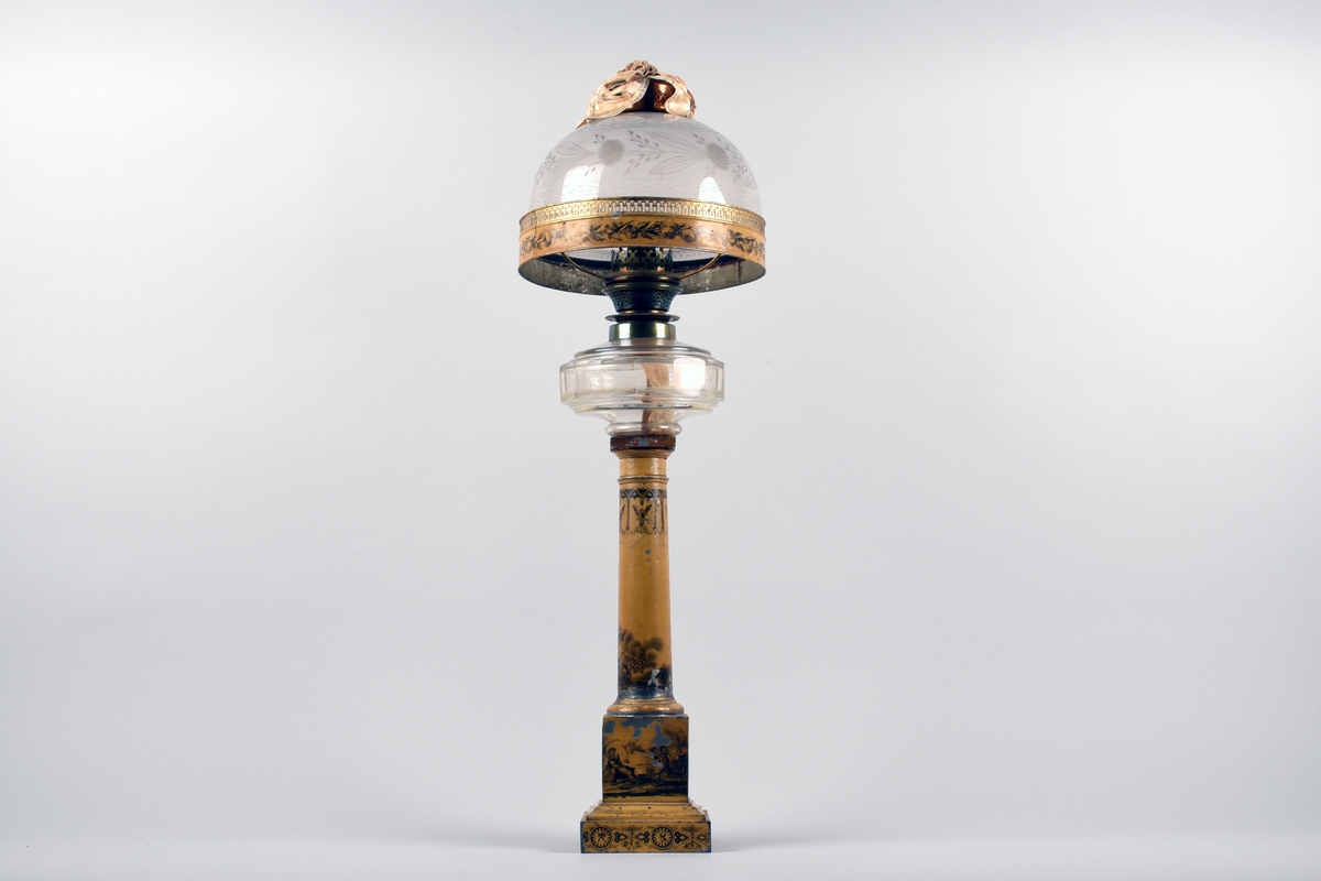 En av ett par fotogenlampor med gulmålad fot i plåt. På målningen finns tryckta stridsscener samt dekor med gotiska valv i svart. Foten är i form av en pelare på kvadratisk bas. Fotogenbehållaren är av glas och brännaren av mässing. Lampkupan är av opalfärgat glas med inslipad blomsterdekor. Högst upp ett skydd sytt i cremefärgat sidentyg, i form av en blomma.