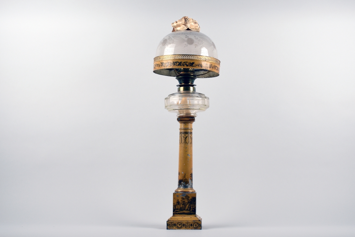 En av ett par fotogenlampor med gulmålad fot i plåt. På målningen finns tryckta stridsscener samt dekor med gotiska valv i svart. Foten är i form av en pelare på kvadratisk bas. Fotogenbehållaren är av glas och brännaren av mässing. Lampkupan är av opalfärgat glas med inslipad blomsterdekor. Högst upp ett skydd sytt i cremefärgat sidentyg, i form av en blomma.