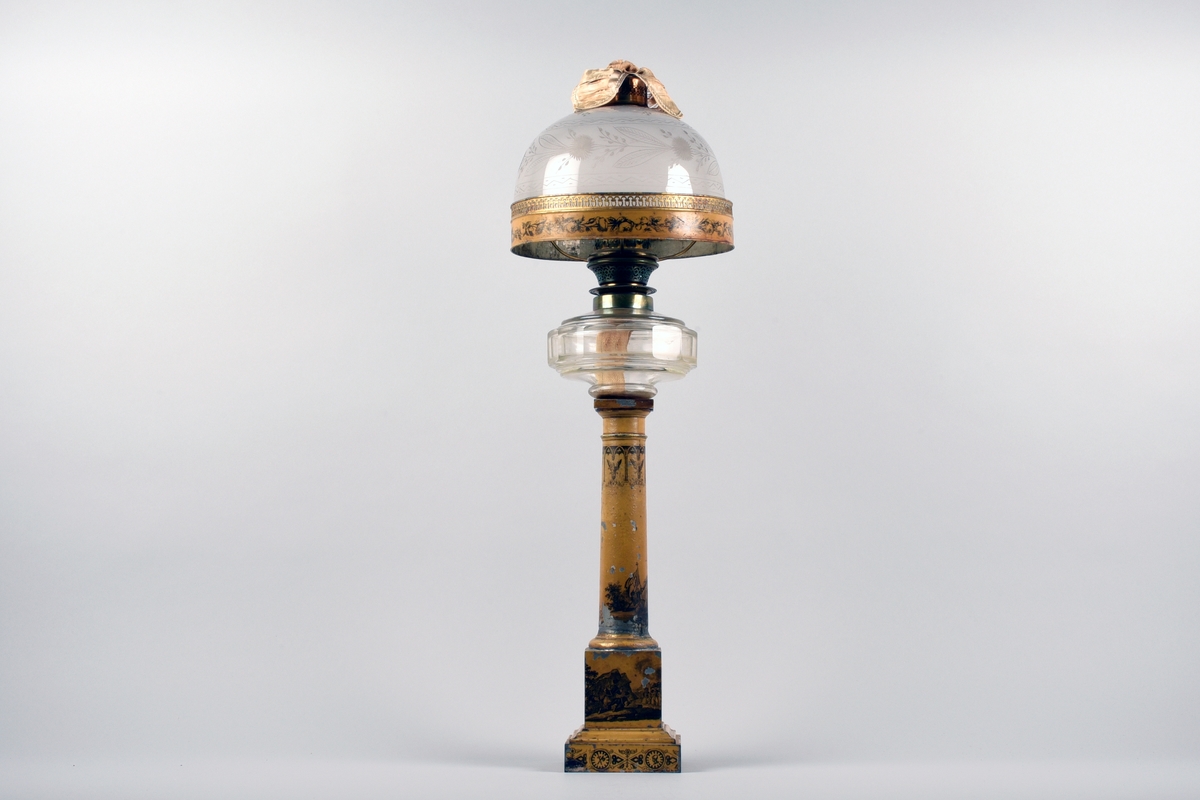 En av ett par fotogenlampor med gulmålad fot i plåt. På målningen finns tryckta stridsscener samt dekor med gotiska valv i svart. Foten är i form av en pelare på kvadratisk bas. Fotogenbehållaren är av glas och brännaren av mässing. Lampkupan är av opalfärgat glas med inslipad blomsterdekor. Högst upp ett skydd sytt i cremefärgat sidentyg, i form av en blomma.