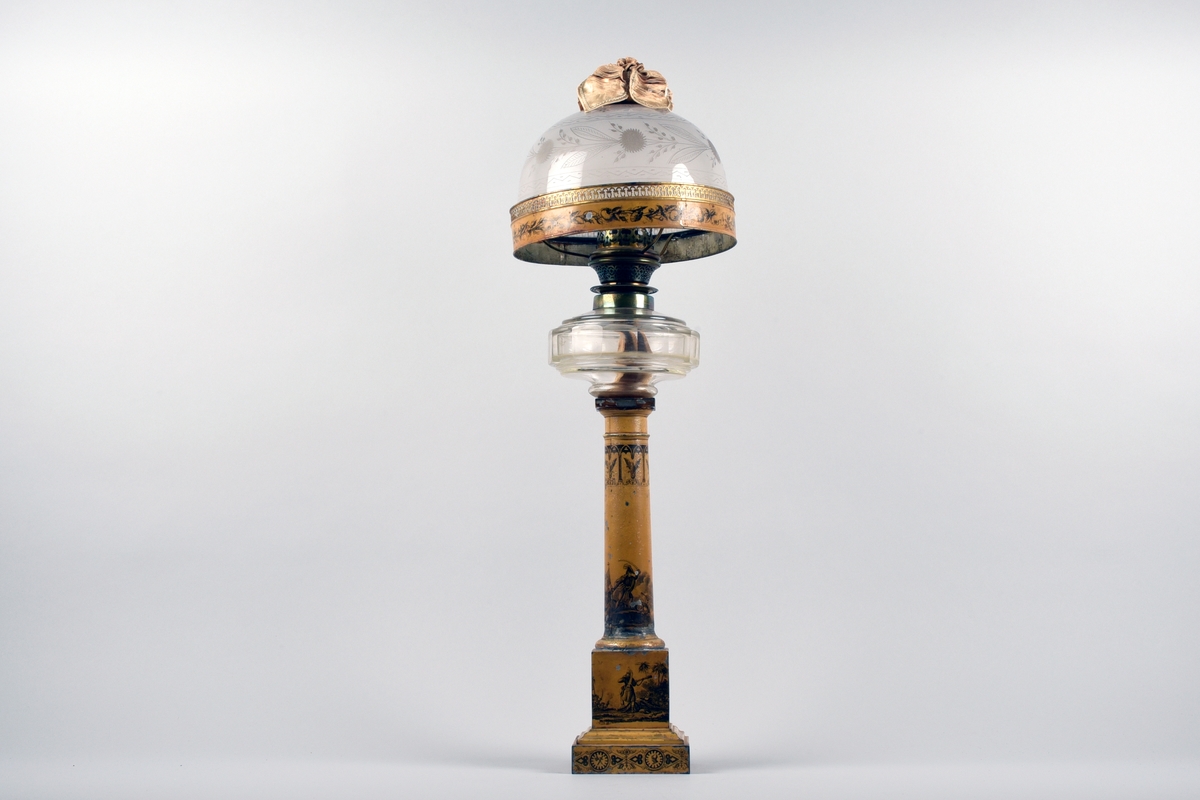 En av ett par fotogenlampor med gulmålad fot i plåt. På målningen finns tryckta stridsscener samt dekor med gotiska valv i svart. Foten är i form av en pelare på kvadratisk bas. Fotogenbehållaren är av glas och brännaren av mässing. Lampkupan är av opalfärgat glas med inslipad blomsterdekor. Högst upp ett skydd sytt i cremefärgat sidentyg, i form av en blomma.