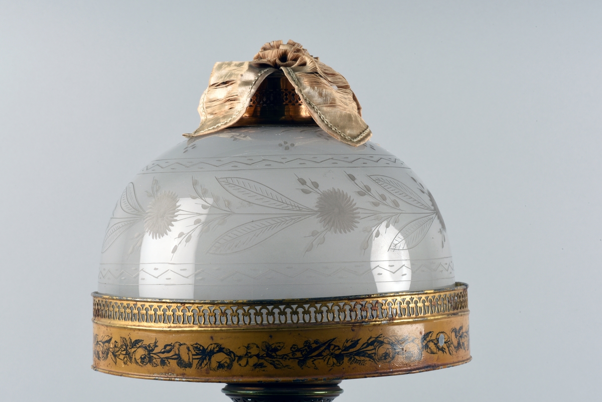 En av ett par fotogenlampor med gulmålad fot i plåt. På målningen finns tryckta stridsscener samt dekor med gotiska valv i svart. Foten är i form av en pelare på kvadratisk bas. Fotogenbehållaren är av glas och brännaren av mässing. Lampkupan är av opalfärgat glas med inslipad blomsterdekor. Högst upp ett skydd sytt i cremefärgat sidentyg, i form av en blomma.