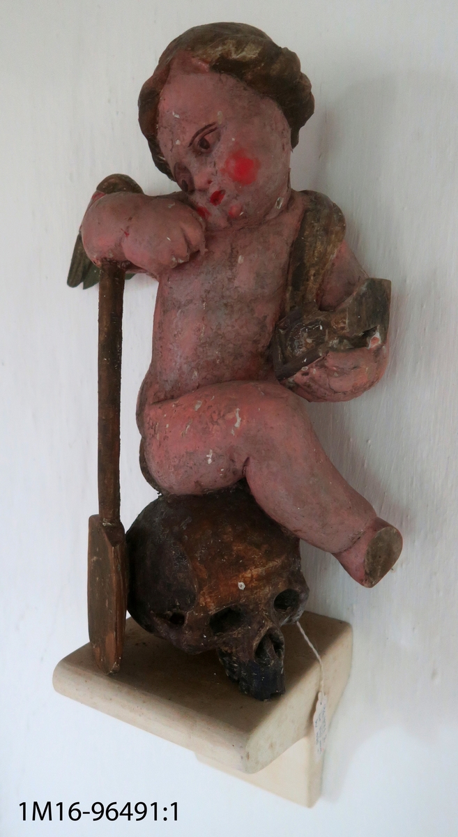 Skulpterad ängel av trä. Sitter på dödskalle med huvudet något högerböjt. Håller en snäcka med tre frukter (?) i höger hand. Draperat tygstycke runt midjan, över höger axel och rygg, avslutas framtill. Saknar vingar, utsträckningar i skuldrorna visar var de suttit. Vänster arm och höger fot saknas. Har hållit ett nu avbrutet föremål i munnen. Träplugg i botten avbruten. Sekundär målning i mörk hudfärg med röda kinder. Hår, dödskalle och övriga detaljer i brunt. Färgen fläckvis borta. Har troligen tillhört epitafium eller altaruppsats.