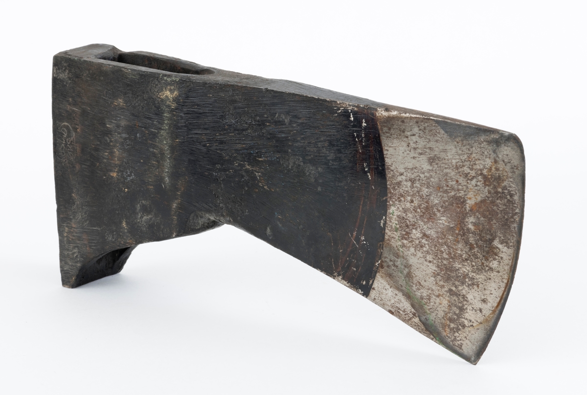 Uskjeftet øks fra produsenten O. Mustad & Søn på Gjøvik i Oppland. Øksehodet er 21,1 centimeter høyt, målt vinkelrett fra midten av den noenlunde plane nakkeflata ned mot den fremre delen av egglinja. Den nederste delen av øksebladet er 9,0 centimeter bred. Her er målet tatt i rett linje mellom den konvekse eggens ytterkanter. Den fremre tverrenden på øksehodet er noenlunde rettlinjet. På «baksida» skrår bladet noe innover mot et punkt like under «øyet» (skafthullet), der bredden er 5,7 centimeter. På denne sida øker bredden på «kjakene» mot den bakre enden av nakkepartiet. Hammerflata på denne delen av øksa er 9,6 centimeter langt og om lag 3,1 centimeter bredt bakerst, 2,8 i den fremre enden. På den høyre sida av øksenakken (sett i øksebrukerens perspektiv) finnes et svakt preget ovalt stempel med teksten «MUSTAD ØKS». Den øvre delen av øksehodet er svartlakkert, mens ei cirka 5,5 centimeter bred sone nederst på bladet er stålblank.