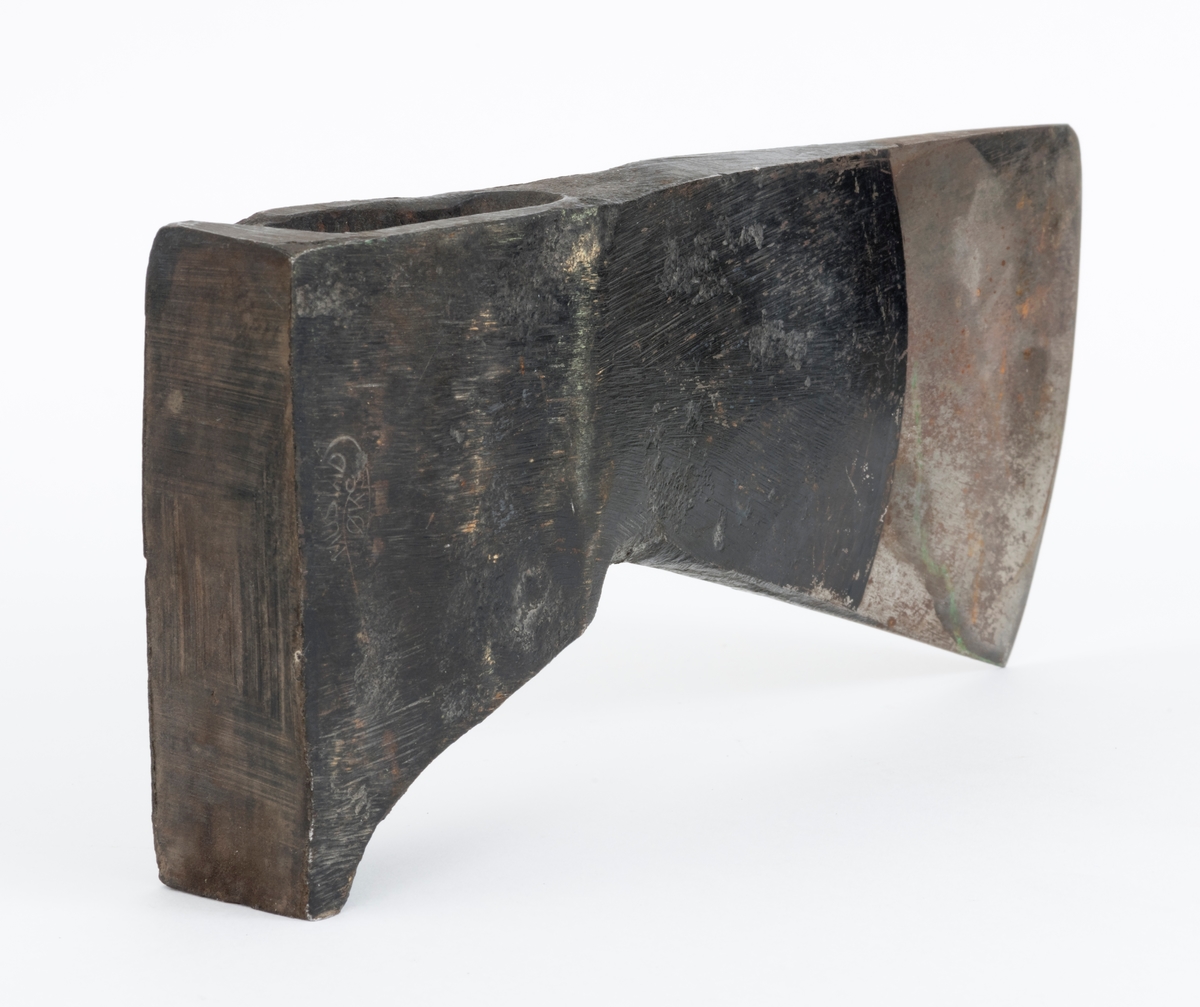 Uskjeftet øks fra produsenten O. Mustad & Søn på Gjøvik i Oppland. Øksehodet er 21,1 centimeter høyt, målt vinkelrett fra midten av den noenlunde plane nakkeflata ned mot den fremre delen av egglinja. Den nederste delen av øksebladet er 9,0 centimeter bred. Her er målet tatt i rett linje mellom den konvekse eggens ytterkanter. Den fremre tverrenden på øksehodet er noenlunde rettlinjet. På «baksida» skrår bladet noe innover mot et punkt like under «øyet» (skafthullet), der bredden er 5,7 centimeter. På denne sida øker bredden på «kjakene» mot den bakre enden av nakkepartiet. Hammerflata på denne delen av øksa er 9,6 centimeter langt og om lag 3,1 centimeter bredt bakerst, 2,8 i den fremre enden. På den høyre sida av øksenakken (sett i øksebrukerens perspektiv) finnes et svakt preget ovalt stempel med teksten «MUSTAD ØKS». Den øvre delen av øksehodet er svartlakkert, mens ei cirka 5,5 centimeter bred sone nederst på bladet er stålblank.