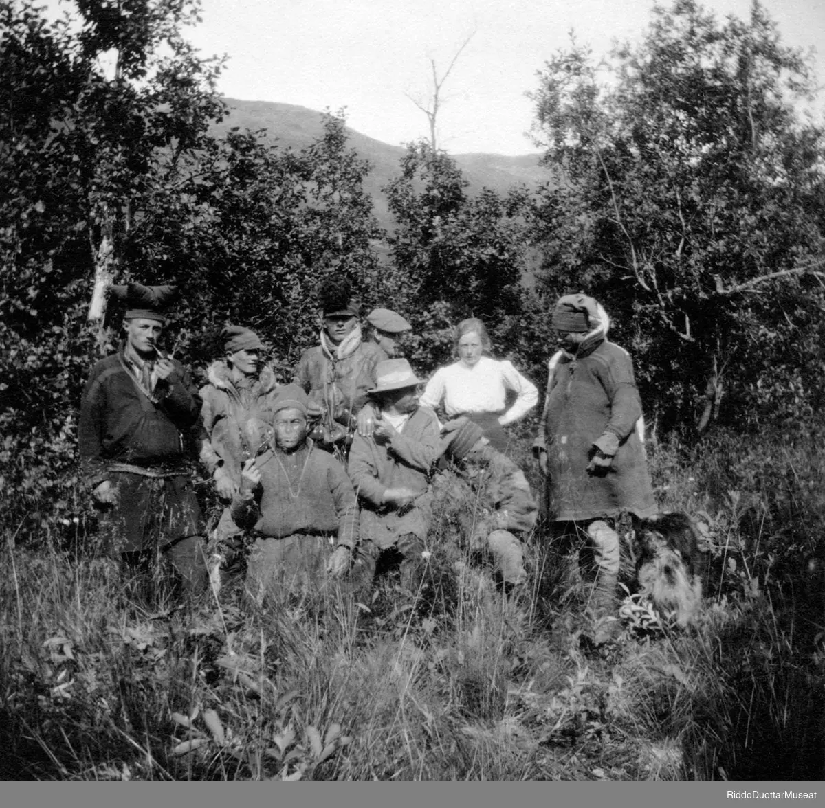 Čieža dievdoolbmo ja okta nissonolmmoš čužžot meahcis.
Syv menn og en kvinne stående i skogen.