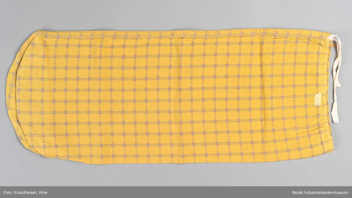 Tekstilpose. Det er søm i den ene siden av posen. Sømmen er en indresøm, det vil si ingen synlige falder og ikke noe stoff som kan trevles opp eller løse tråder på innsiden av posen. Bunnen er sydd av et rundt stoffstykke. Den er festet til posen med en vanlig søm, og kantene er kastet over for hånd. Det er løpegang oppe med bendelbånd gjennom. Dette kan ha vært et trekk til en pute, for eksempel en nakkepute, en pute som ligger i kortendene på en sofa eller seng som res opp om dagen. Det sitter en merkelapp på innsiden av sømmen i øverste kant. Det er en hvit lapp med rød skrift (maskinbroderi). Elektriker. Ovenfor dette merket er det sydd noen få, ujevne sting med beige brodergarn. Dette kunne ha vært en reparasjon, men det er ingen synlige skader i stoffet som skulle tilsi noe slikt. Det kan også være en vaskerimerking, eventuelt rester av en vaskerimerking. Det er også feste en liten, oransje tråd i kanten oppe. Det finnes noe liknende på andre av tekstilene – noe som kan tyde på at det er en merking som sier noe om avdelingen tekstilet hører til – eller det kan være en vaskerimerking. Det er søm i den ene siden av posen.