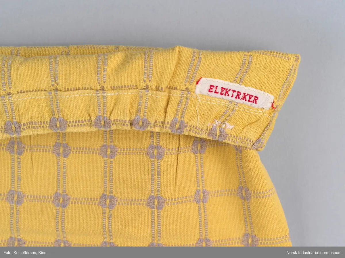 Tekstilpose. Det er søm i den ene siden av posen. Sømmen er en indresøm, det vil si ingen synlige falder og ikke noe stoff som kan trevles opp eller løse tråder på innsiden av posen. Bunnen er sydd av et rundt stoffstykke. Den er festet til posen med en vanlig søm, og kantene er kastet over for hånd. Det er løpegang oppe med bendelbånd gjennom. Dette kan ha vært et trekk til en pute, for eksempel en nakkepute, en pute som ligger i kortendene på en sofa eller seng som res opp om dagen. Det sitter en merkelapp på innsiden av sømmen i øverste kant. Det er en hvit lapp med rød skrift (maskinbroderi). Elektriker. Ovenfor dette merket er det sydd noen få, ujevne sting med beige brodergarn. Dette kunne ha vært en reparasjon, men det er ingen synlige skader i stoffet som skulle tilsi noe slikt. Det kan også være en vaskerimerking, eventuelt rester av en vaskerimerking. Det er også feste en liten, oransje tråd i kanten oppe. Det finnes noe liknende på andre av tekstilene – noe som kan tyde på at det er en merking som sier noe om avdelingen tekstilet hører til – eller det kan være en vaskerimerking. Det er søm i den ene siden av posen.