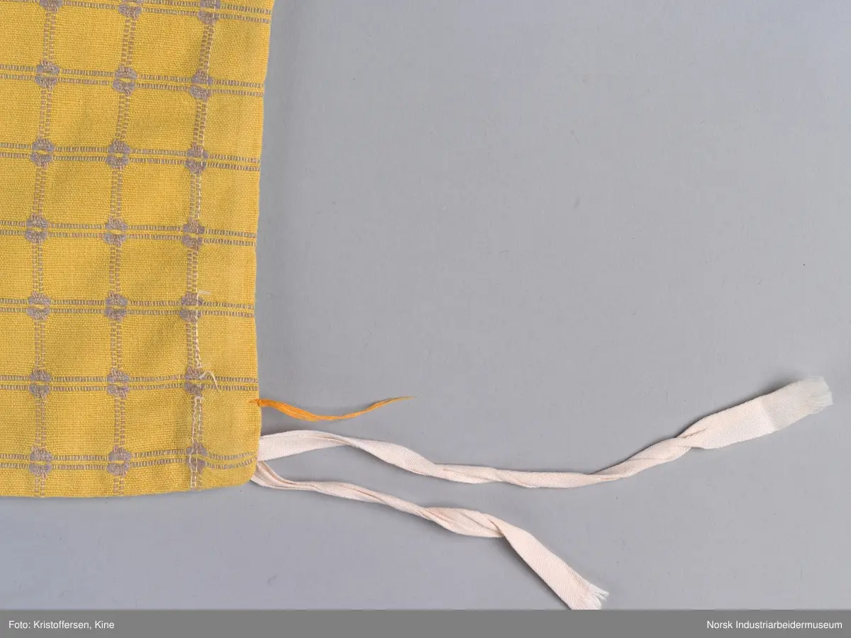 Tekstilpose. Det er søm i den ene siden av posen. Sømmen er en indresøm, det vil si ingen synlige falder og ikke noe stoff som kan trevles opp eller løse tråder på innsiden av posen. Bunnen er sydd av et rundt stoffstykke. Den er festet til posen med en vanlig søm, og kantene er kastet over for hånd. Det er løpegang oppe med bendelbånd gjennom. Dette kan ha vært et trekk til en pute, for eksempel en nakkepute, en pute som ligger i kortendene på en sofa eller seng som res opp om dagen. Det sitter en merkelapp på innsiden av sømmen i øverste kant. Det er en hvit lapp med rød skrift (maskinbroderi). Elektriker. Ovenfor dette merket er det sydd noen få, ujevne sting med beige brodergarn. Dette kunne ha vært en reparasjon, men det er ingen synlige skader i stoffet som skulle tilsi noe slikt. Det kan også være en vaskerimerking, eventuelt rester av en vaskerimerking. Det er også feste en liten, oransje tråd i kanten oppe. Det finnes noe liknende på andre av tekstilene – noe som kan tyde på at det er en merking som sier noe om avdelingen tekstilet hører til – eller det kan være en vaskerimerking. Det er søm i den ene siden av posen.