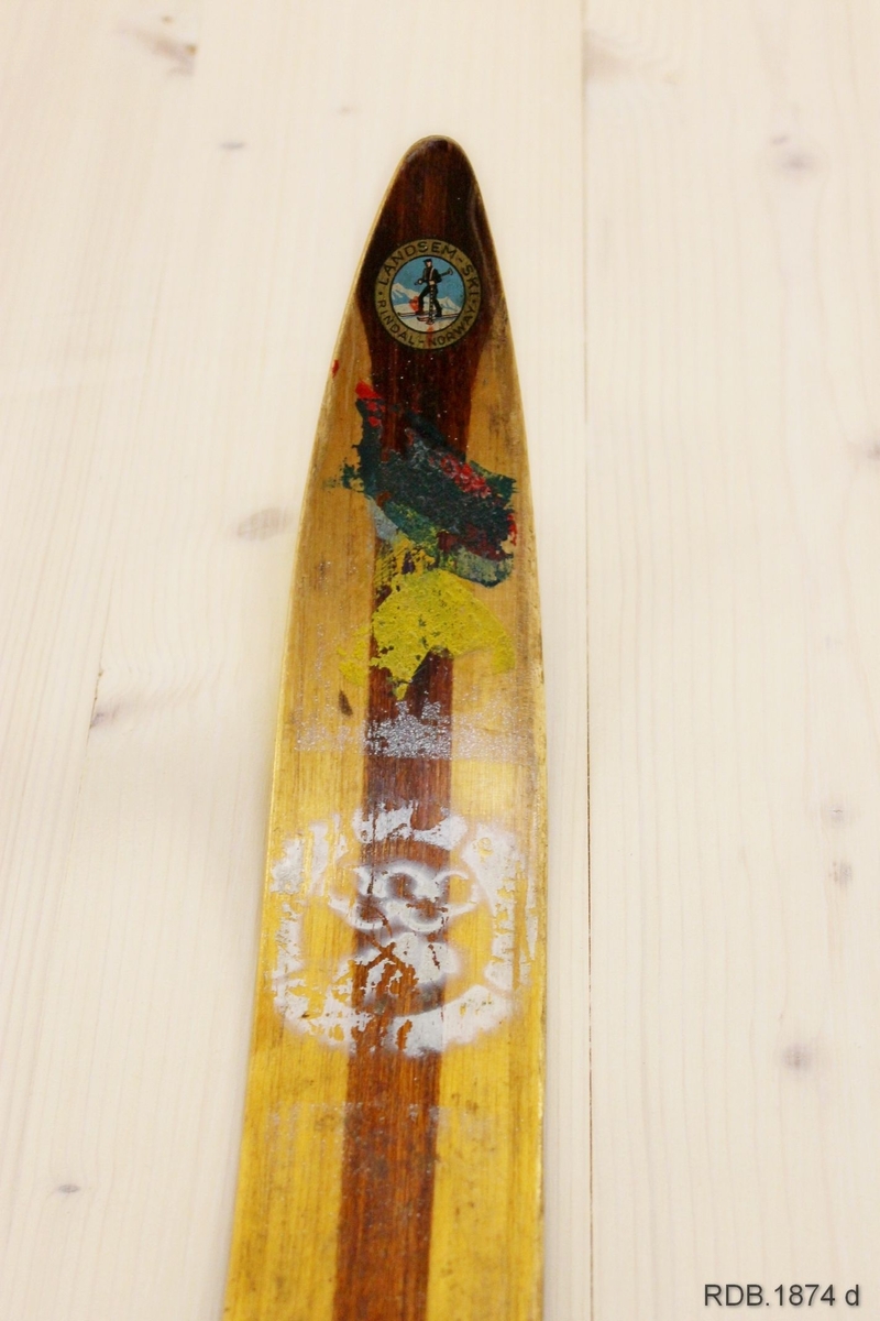 Avsmalende, jevnbred ski med liten bue. Bøy uten tupp. Rett avskåret bakende.
Dekorert med ei brun stripe på trehvit bunn.

Noen stempel fra OL øverst på skiet.