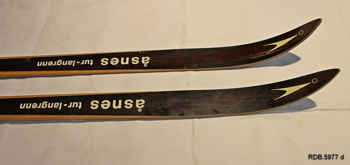 Et par brune, laminerte treski med trekvite kanter, kvit påskrift og dekor. Åsnes-logo på skituppen. Hickorysåle og lignostonekanter. Smal skitupp og stålkanter bak. Påmontert gullfarga rottefellabindinger og to kvite fotplater. Bruksanvisning bak bindingen.