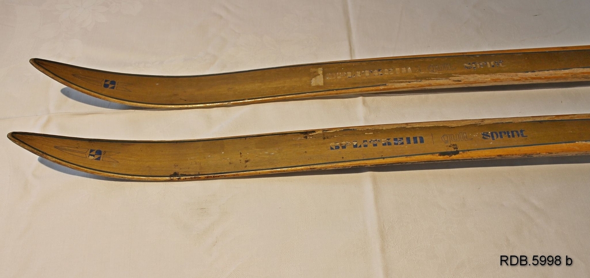 Et par laminerte treski. Trekvite med et bredt gullfarga felt kanta med en smal blå stripe langsetter skiet. Blå Splitkeinlogo på skituppen og blå påskrift. Påmontert gullfarga Rottefellabindinger. Bak disse er faststifta to kvite fotplater, den bakerste har metallkanter.